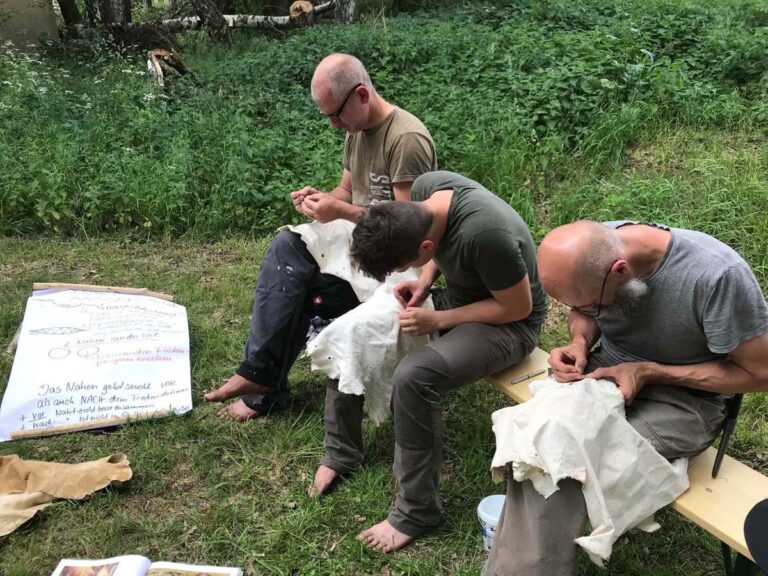 Drei Männer nähen draußen auf Bänken.