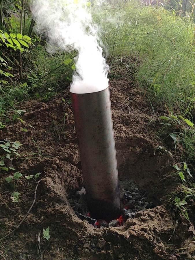 Rauchender Metallrohr im Boden, umgeben von Grün.