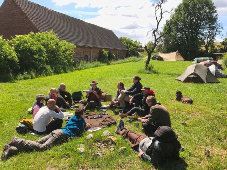 Menschen sitzen im Freien im Kreis, Zelte im Hintergrund.